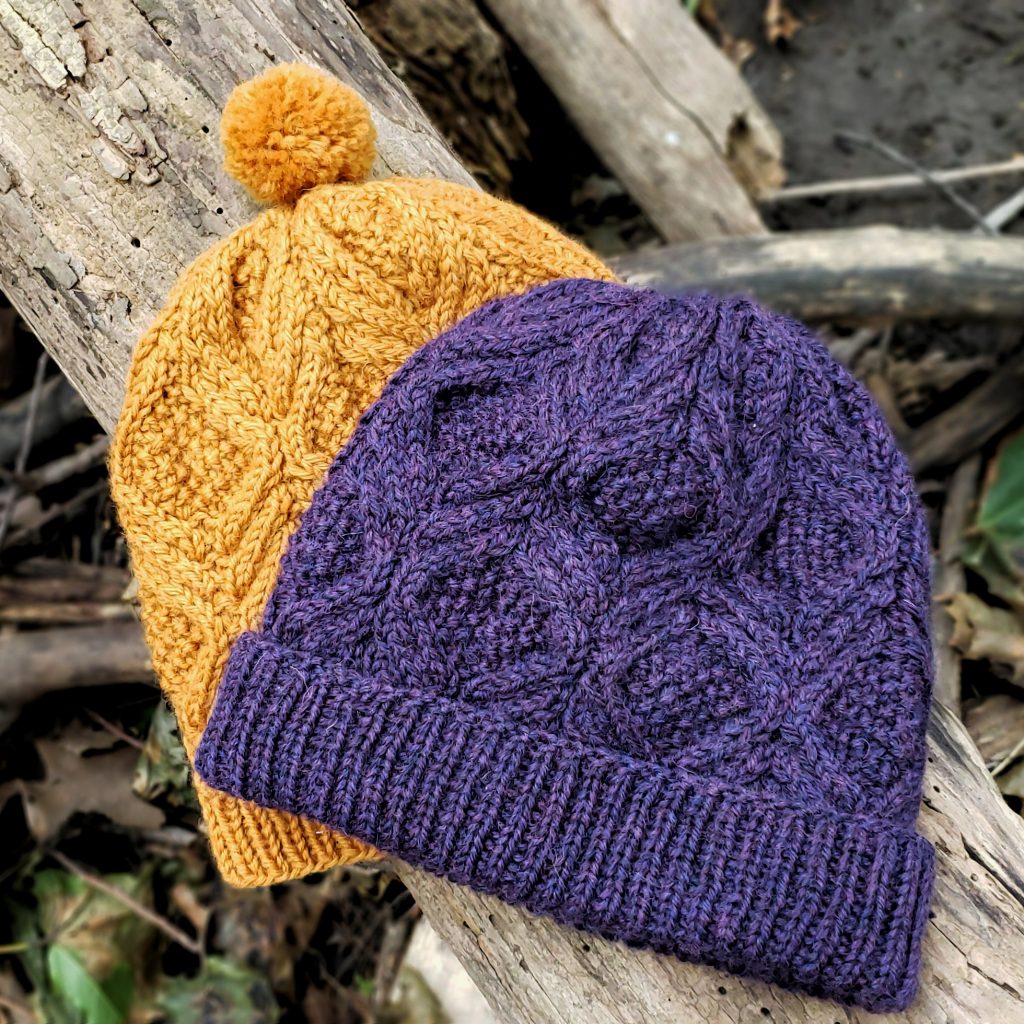 Two cabled hats, the same pattern but one yellow, one purple, laid on a log.